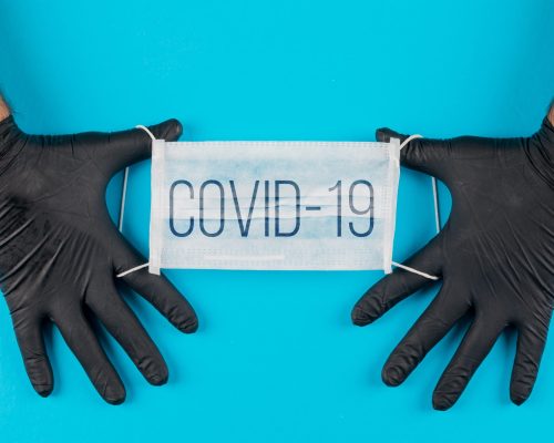 Medical mask with text covid-19 in a hands with black gloves top view on a cyan blue background